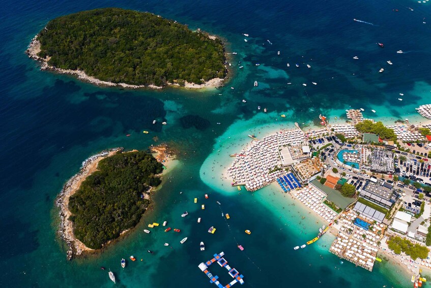 An aerial view of a coastal area with clear turquoise waters, a densely populated beach with numerous umbrellas and sunbeds, and several small islands with rocky coastlines. 3