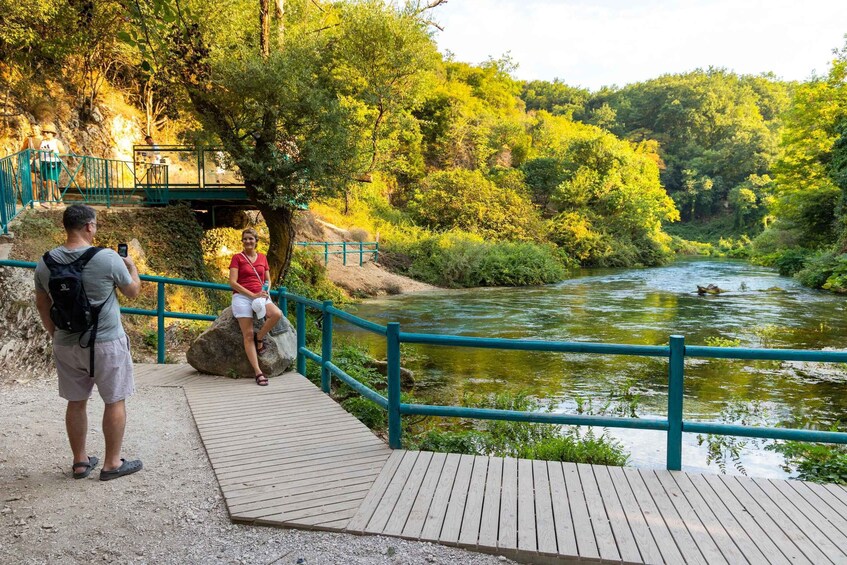 A wooden boardwalk with a metal railing alongside a river, surrounded by lush greenery and trees. 5