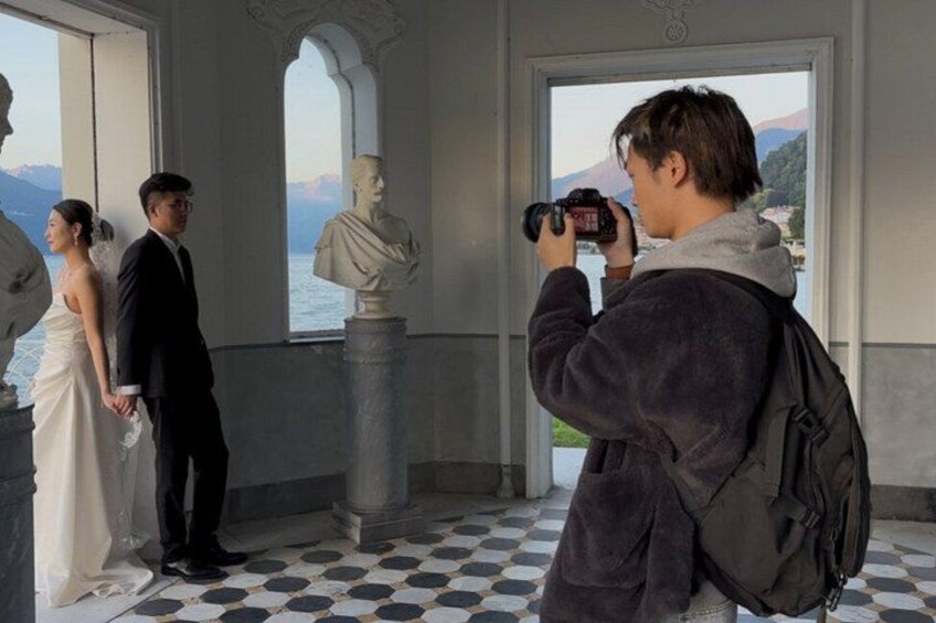 A couple in wedding attire, a photographer, and a bust sculpture in a room with large windows. 1
