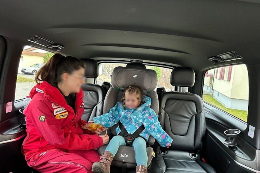 A woman in a red jacket and a child in a blue jacket seated in a car's back seat. 5