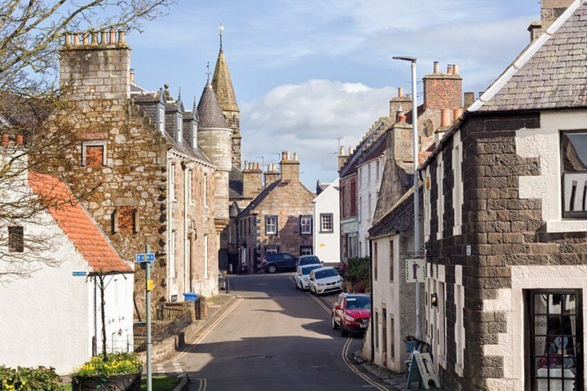 A narrow street lined with historic stone buildings, a church tower, and parked cars. 4