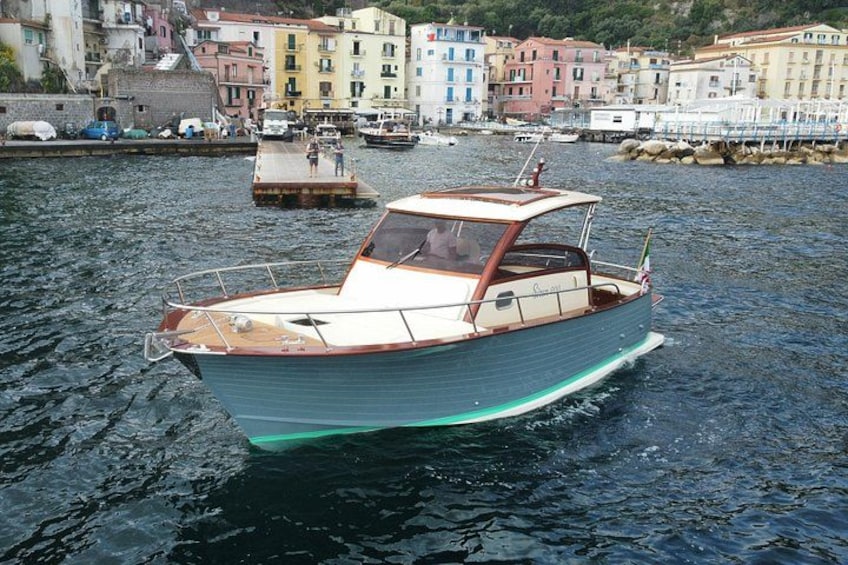 A narrowboat on calm water with a dock in the background and colorful buildings along the shore. 1