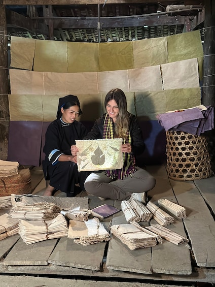 Two people sitting on a mat, holding a patterned cloth, with stacks of dried bamboo and woven baskets around them. 2