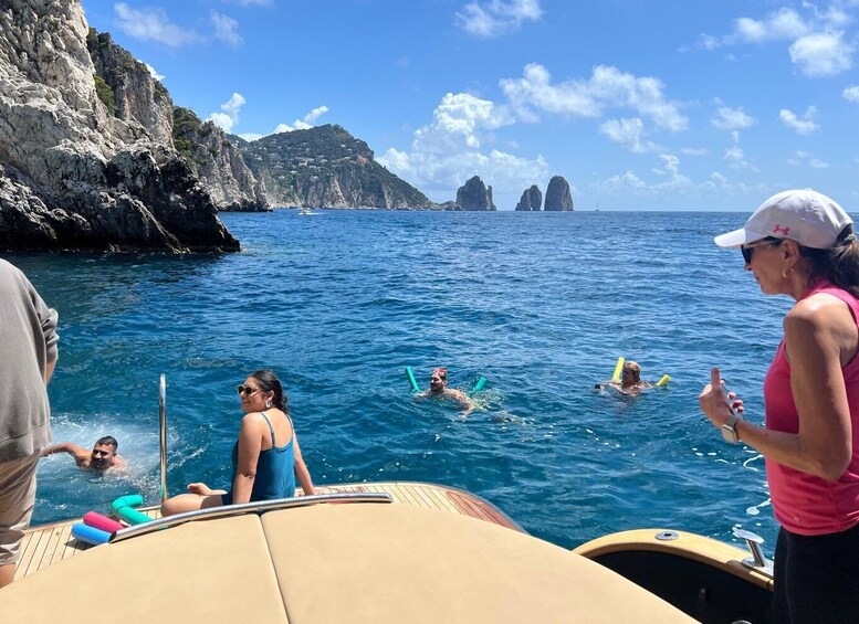 A group of people swimming and floating on a boat in a coastal area with rocky cliffs and clear blue water. 3