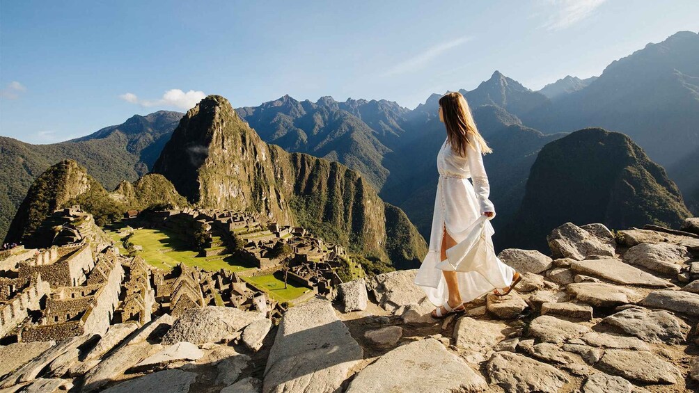 A woman in a white dress standing on rocky terrain with Machu Picchu ruins in the background. 3