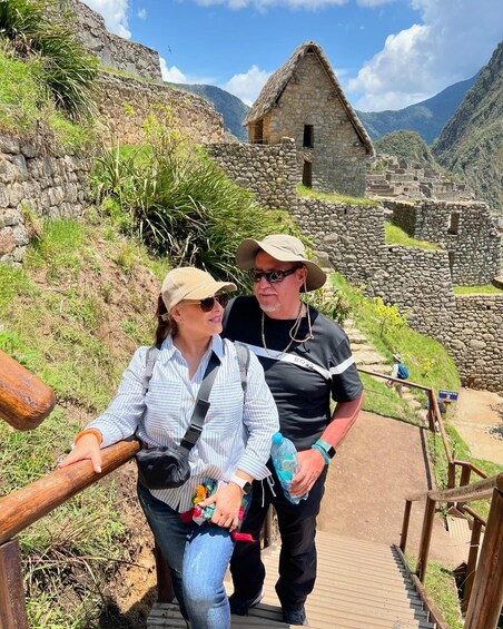 Two people standing on a wooden staircase with a stone building and a thatched-roof structure in the background. 2