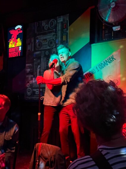 A stage with two people, one holding a microphone, in a dimly lit venue with a colorful backdrop and a visible logo. 2