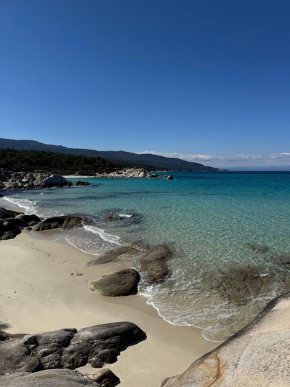 En strand med klart turkis vand, en klippefyldt kystlinje og et fjerntliggende bjerglandskab. 3