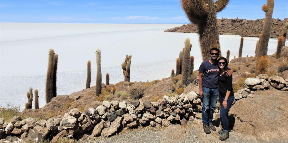 A couple standing on rocky terrain with cacti and a body of water in the background. 5
