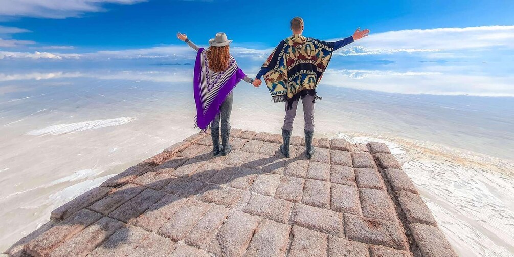 Two people standing on a stone platform with their arms outstretched, overlooking a vast salt flat under a clear blue sky. 4