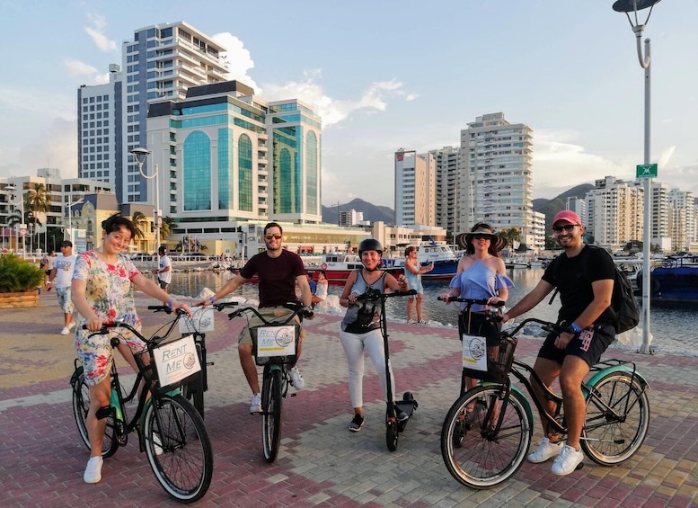 A group of people on bicycles with 'RENT ME' signs, by a waterfront with buildings and boats in the background. 1