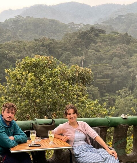 Twee personen zitten aan een houten tafel buiten, met een weelderig groen bos op de achtergrond. 2