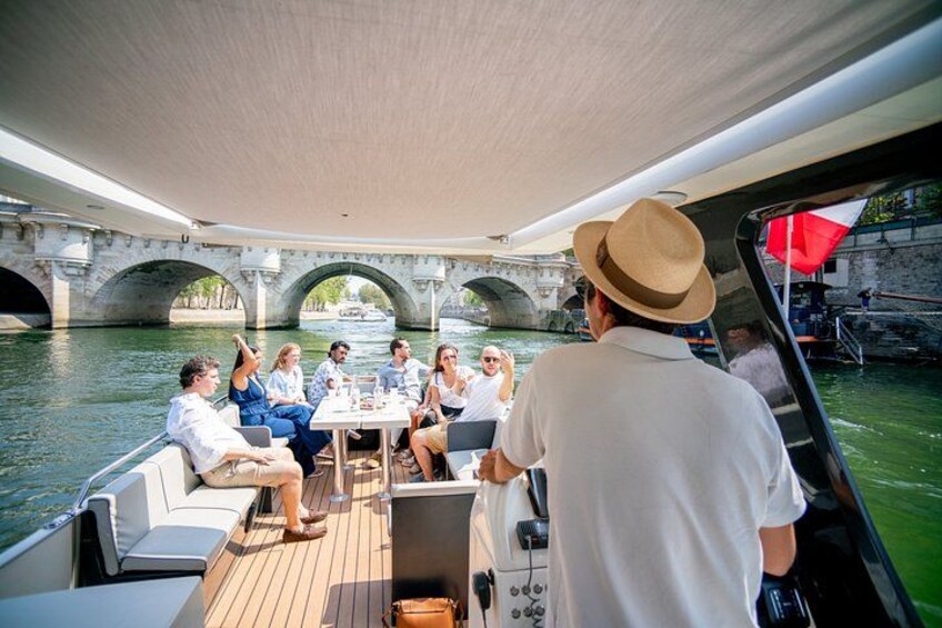 A group of people on a boat with a table, a man in a hat, and a bridge in the background. 1