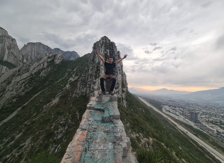 A person standing on a narrow stone path with arms raised, surrounded by steep mountains and a distant cityscape. 3
