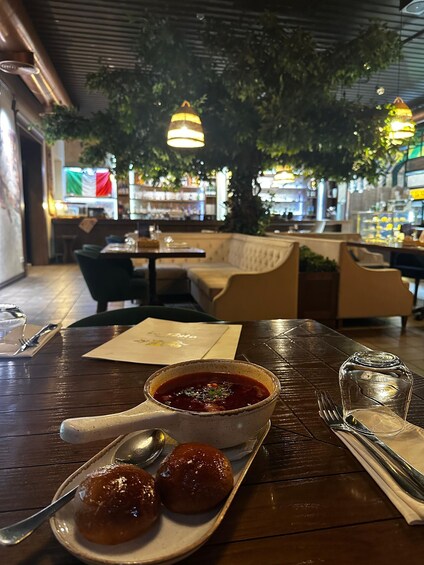 A restaurant setting with a table set for two, featuring a bowl of soup, two bread rolls, and cutlery. 2