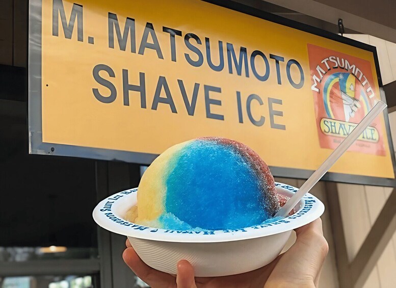 A person holding a bowl of shaved ice with a spoon, in front of a sign that reads 'M. Matsumoto Shave Ice'. 4