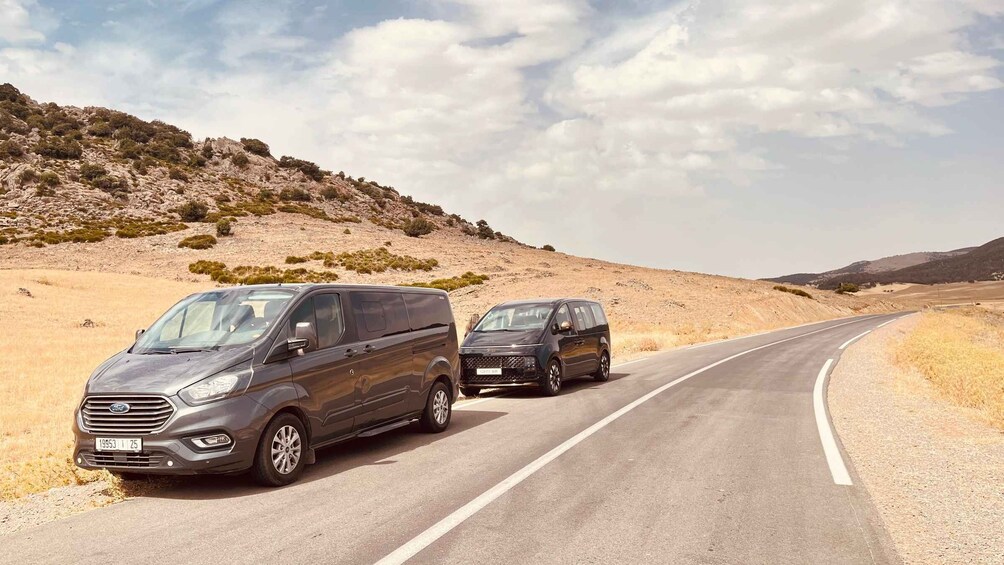 Two dark-colored vans parked on a winding road in a desert landscape with sparse vegetation and hills in the background. 1