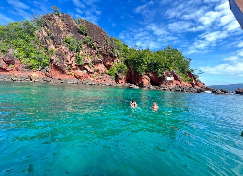 A clear turquoise sea with two people swimming, surrounded by rocky cliffs and lush greenery. 5