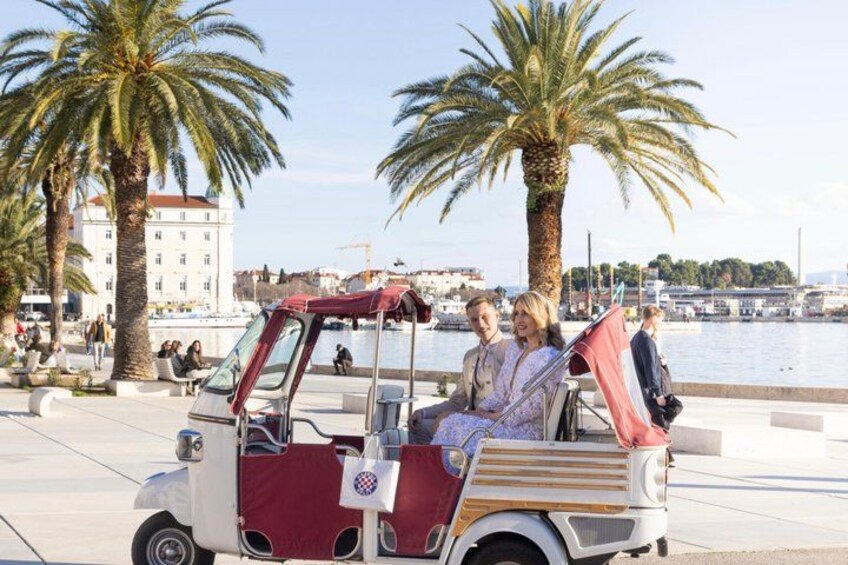 A vintage car with passengers, palm trees, and a waterfront in the background. 1