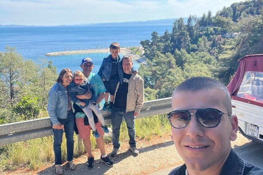 A group of people standing on a roadside with a scenic view of the sea and hills in the background. 2