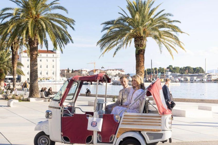 A vintage car with passengers, palm trees, and a waterfront in the background. 1