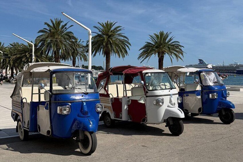 Tre tuk-tuks står parkerade på en solig gata, med palmer och ett kryssningsfartyg i bakgrunden. 2