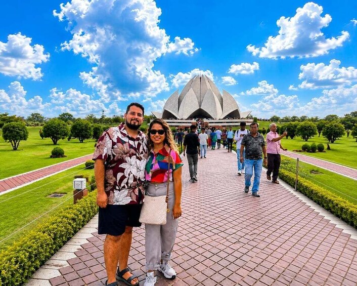 A couple standing in front of a large, modern architectural structure with a clear blue sky and scattered clouds in the background. 3