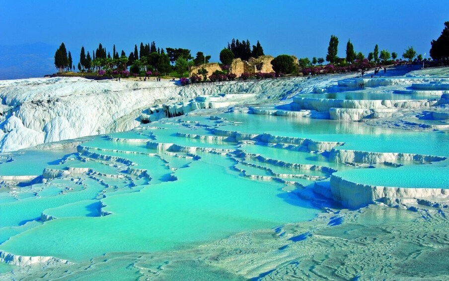 Terraced travertine formations with clear turquoise pools, surrounded by trees and a distant mountain range. 3