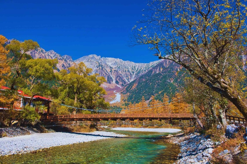 A wooden bridge over a clear river, surrounded by autumn-colored trees and mountains in the background. 1