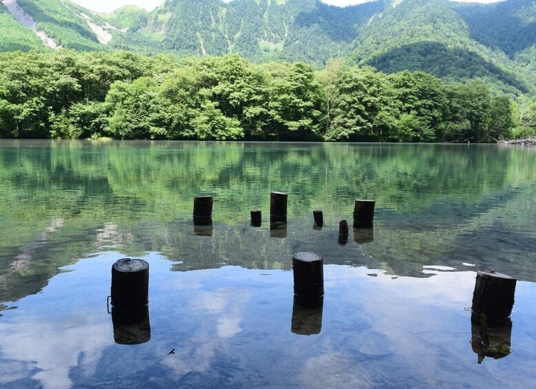 A clear lake with wooden posts in the foreground and a forested mountain range in the background. 1