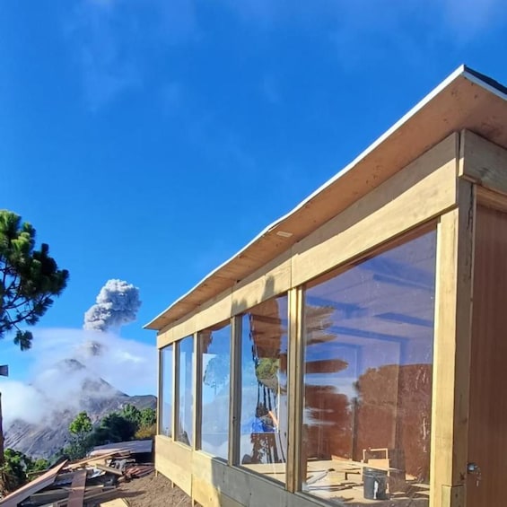 A wooden building with large windows, construction materials, and a mountain with smoke in the background. 1