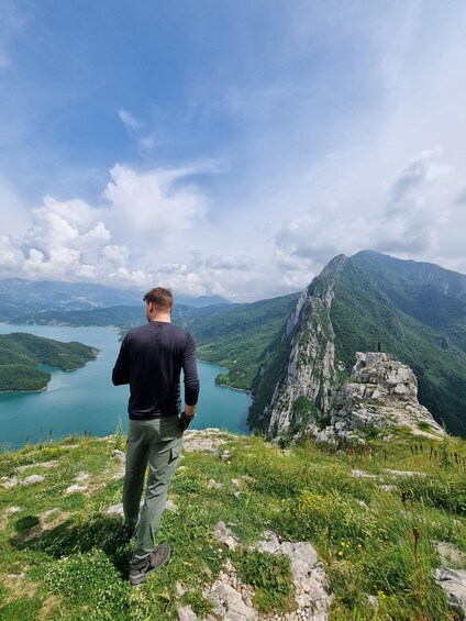 A man standing on a grassy hill overlooking a lake and mountains. 1
