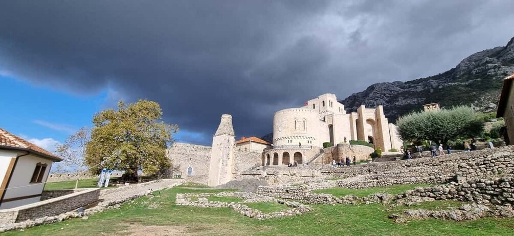 An ancient fortress with stone walls and a tower, surrounded by grassy areas and a few people. 2
