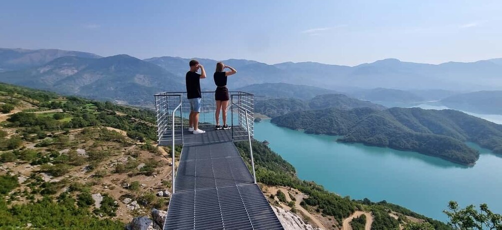 Two people standing on a metal platform overlooking a turquoise lake surrounded by mountains. 3