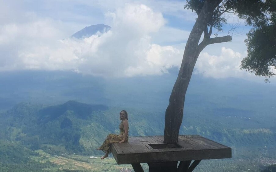 A person sitting on a wooden platform with a tree, overlooking a mountainous landscape. 5