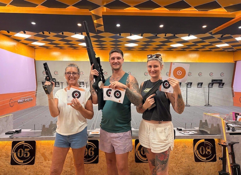 Three people holding firearms and target sheets in an indoor shooting range. 1