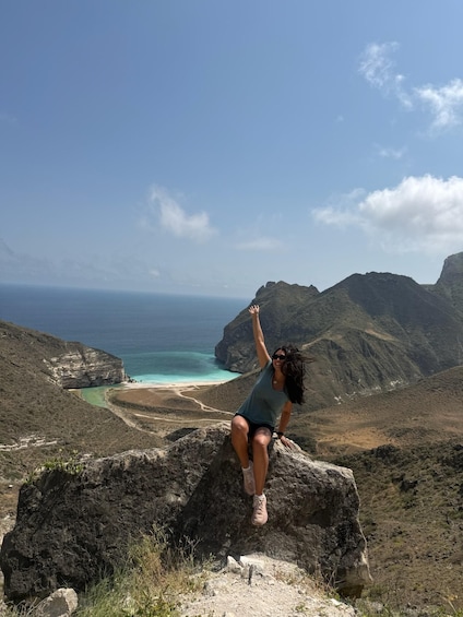 A person sitting on a rock with arms raised, overlooking a coastal landscape with mountains and a beach. 5