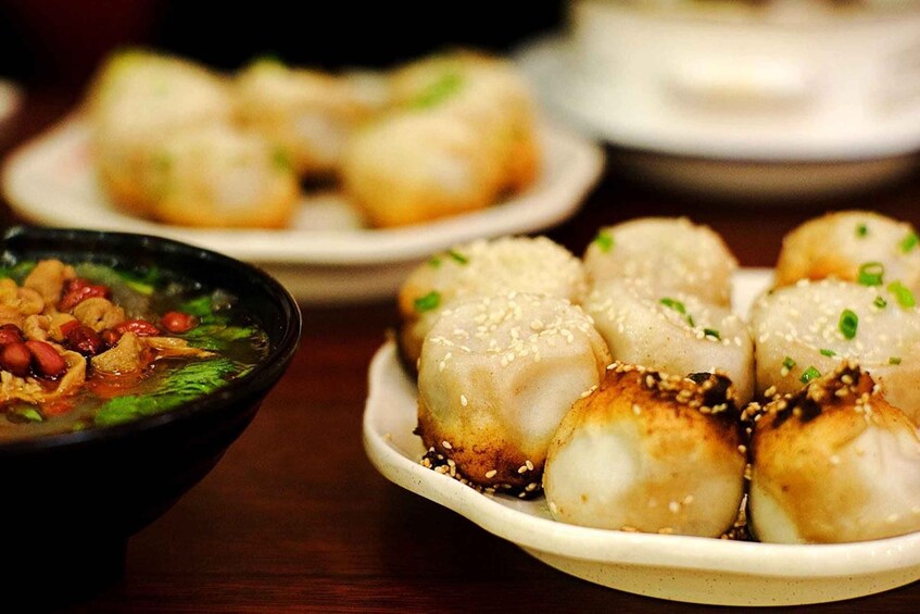 A plate of steamed buns garnished with sesame seeds and chopped green onions, accompanied by a bowl of soup with vegetables and peanuts. 4