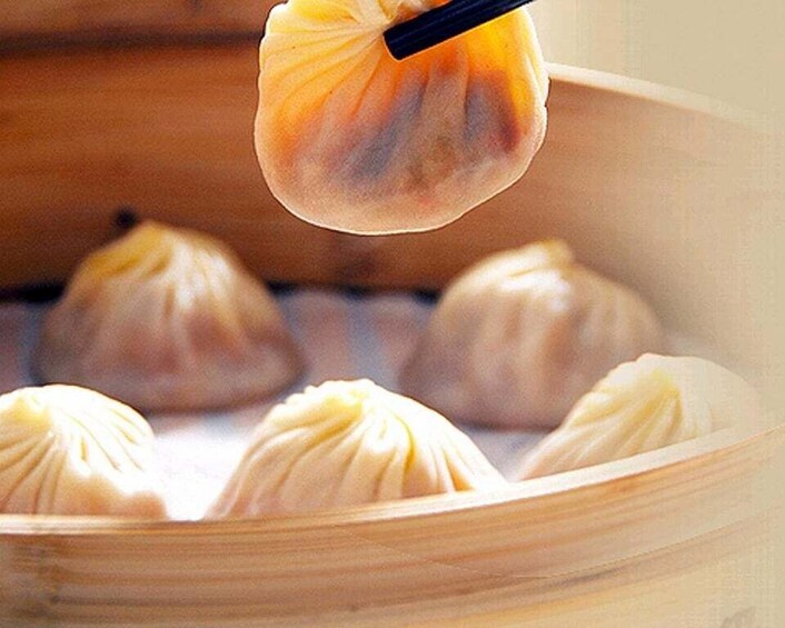 A hand holding a dumpling over a bamboo steamer with several dumplings inside. 1