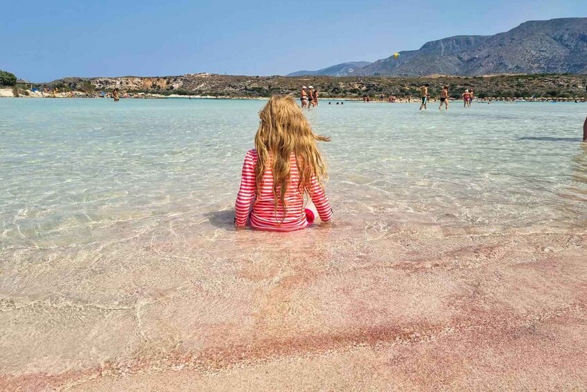 A child sitting in shallow water at a beach with clear skies and mountains in the background. 4