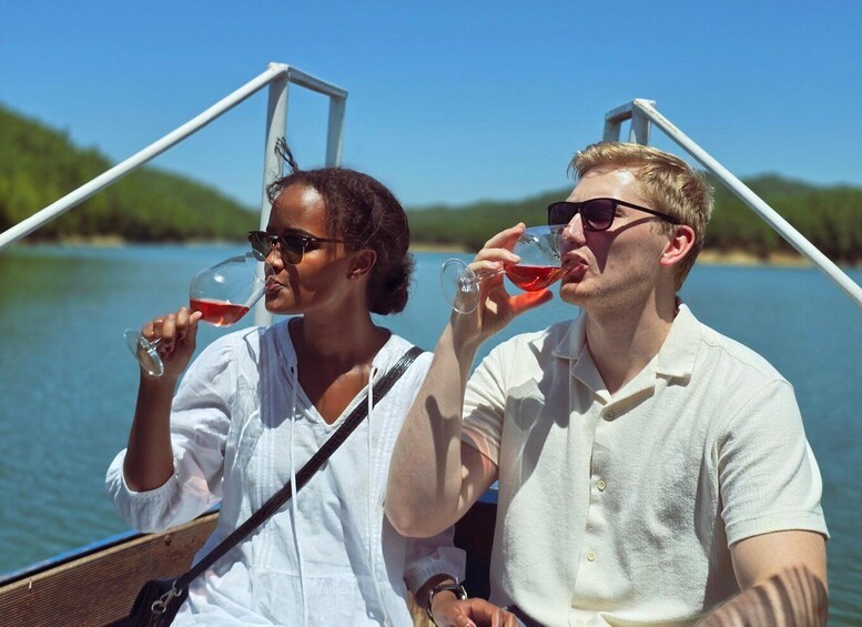 Two people on a boat, each holding a glass of red wine, with a body of water and greenery in the background. 1