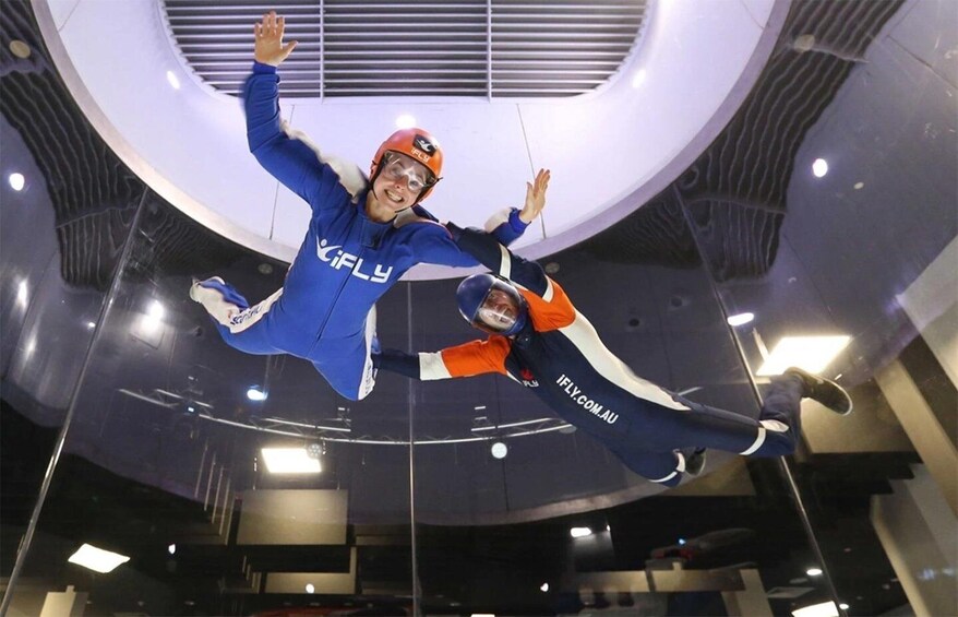 Two people in blue and orange skydiving suits with helmets, suspended in mid-air in a modern indoor facility with circular architecture. 5