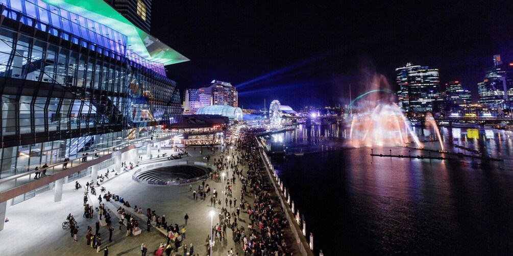 A nighttime cityscape with a waterfront promenade, illuminated buildings, and a water fountain display. 3