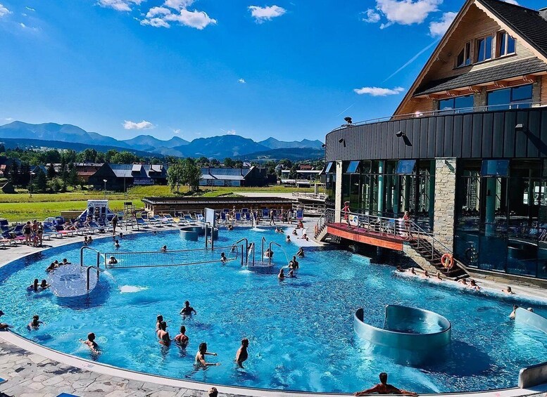 A swimming pool with people, a building, and mountains in the background. 1