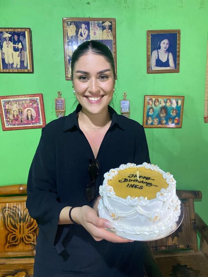 A person holding a birthday cake with "Happy Birthday Jess" written on it, standing in front of a green wall with various framed pictures. 2