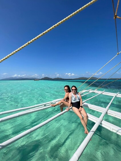 Two people sitting on a boat deck with clear turquoise water and distant islands. 1