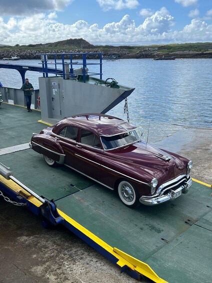 A vintage maroon car parked on a dock by a body of water with a rocky shoreline and hills in the background. 2