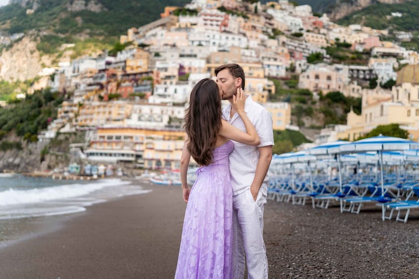 Un couple s’embrasse sur une plage, avec en arrière-plan des bâtiments aux couleurs vives et des parasols bleus. 1