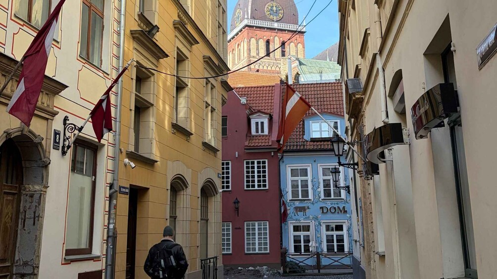 A narrow street with buildings, a clock tower, and flags. 1