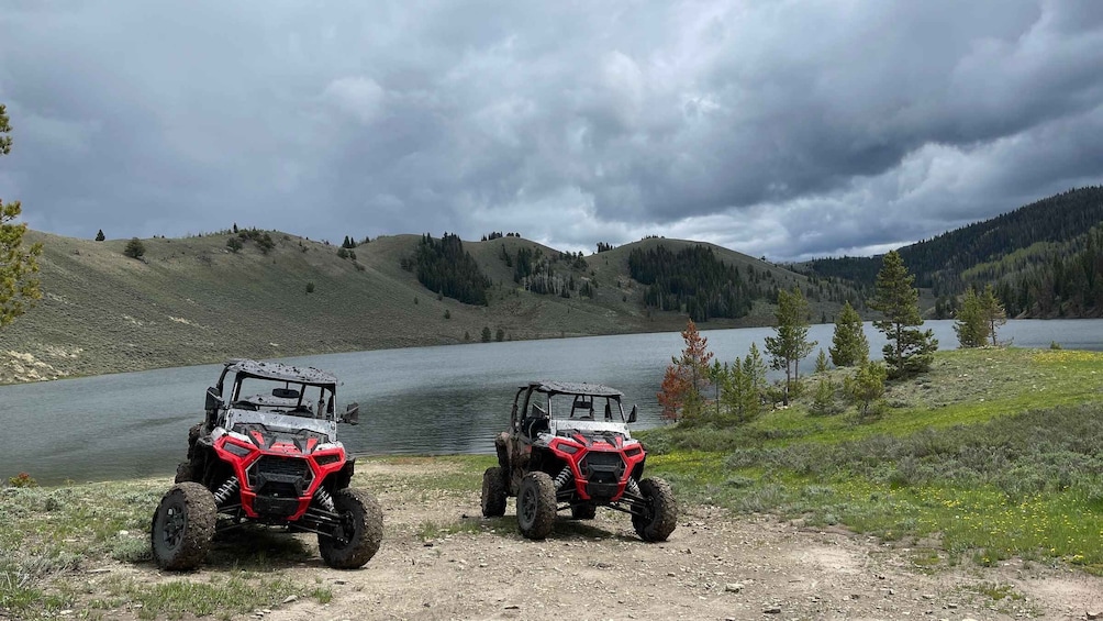 Two red off-road vehicles parked on a dirt path by a lake with hills in the background. 3